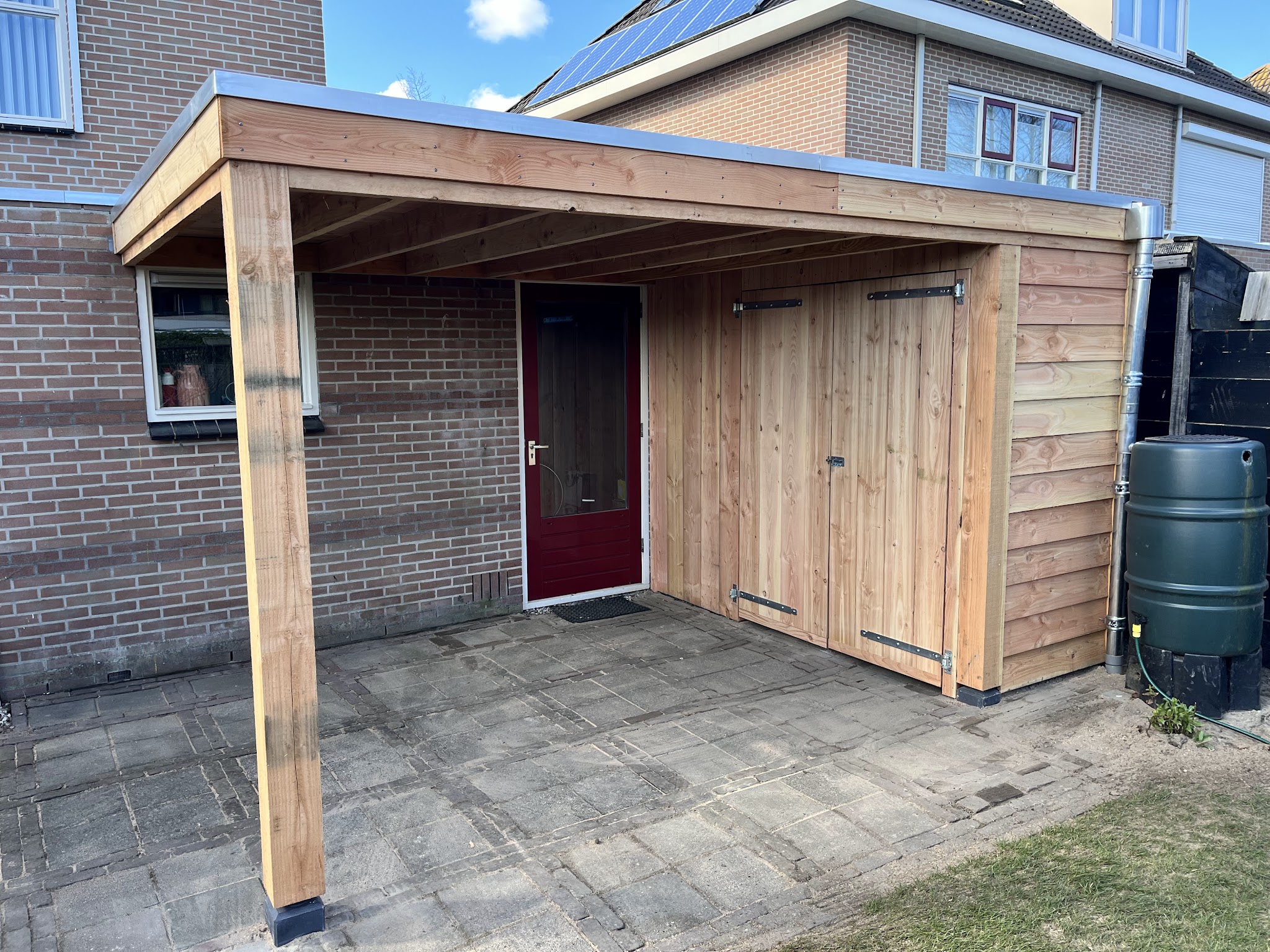 Een nieuwe houten overkapping met berging en dubbele deuren tegen een bakstenen woning